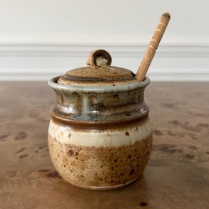 May include: A ceramic honey pot with a speckled brown and cream glaze, featuring a matching lid with a central knob and a wooden honey dipper resting inside. The pot has a rounded body with a flared rim and a smooth, earthy finish throughout.