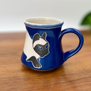 May include: A ceramic mug with a blue exterior and a white interior. The mug features a black and white cat design on the side. The handle is blue, and the mug is sitting on a wooden surface. The mug is a unique piece of drinkware.