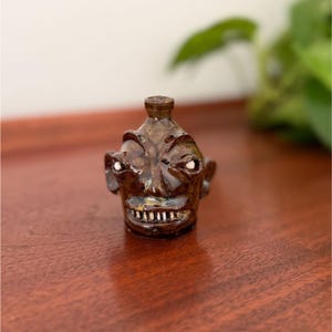 May include: A small, dark brown glazed ceramic jug shaped like a human head with a narrow neck opening at the top. The face features prominent brow ridges, two white eyes, and a wide mouth displaying a row of small, individual white teeth.