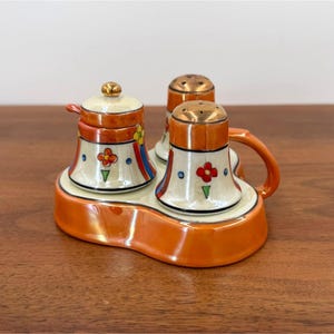 May include: A ceramic condiment set featuring a lidded mustard pot, a salt shaker, and a pepper shaker on a matching orange base. Each piece is cream-colored with orange trim, gold-tone lids, and hand-painted floral motifs in red, yellow, and blue.