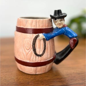 May include: A novelty ceramic mug shaped like a wooden barrel, with a cowboy figure forming the handle. The mug is light pink with dark red bands and a cowboy in a blue shirt and black hat. The cowboy is holding a rope.