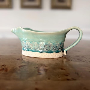 May include: A ceramic gravy boat with a turquoise glaze and a textured, wave-like design. The handle is curved, and the base is a creamy white. The boat is designed for serving sauces and gravies.