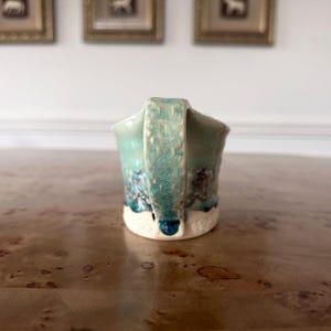 May include: A handmade ceramic mug with a turquoise glaze and a textured handle. The mug features a white base and dark blue accents, creating a unique, artistic design. The mug is perfect for coffee, tea, or any beverage.