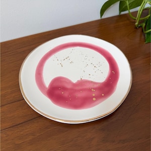 Puede incluir: Un plato de cer&aacute;mica blanco con borde dorado y un dise&ntilde;o abstracto audaz en tonos rosa y dorado. El dise&ntilde;o presenta una forma grande y curvada con motas doradas, creando una est&eacute;tica moderna y art&iacute;stica. El plato est&aacute; sobre una superficie de madera.