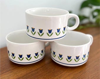 May include: Three white ceramic soup mugs stacked on a wooden surface. Each mug features a blue and green floral pattern around the rim. One mug has a handle. The mugs are stacked in a triangular arrangement.