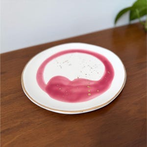 Puede incluir: Un plato de cer&aacute;mica blanco con borde dorado y un dise&ntilde;o abstracto en rosa y dorado. El dise&ntilde;o presenta una forma circular con un elemento en forma de coraz&oacute;n y detalles dorados. El plato est&aacute; sobre una superficie de madera.