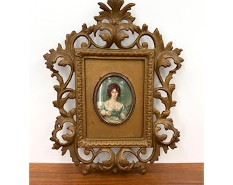 May include: A gold-colored wall frame with an ornate, scrolling leaf design surrounding a rectangular border. Centered within is an oval portrait of a person with reddish-brown hair wearing a light-colored garment and a necklace, set against a background.