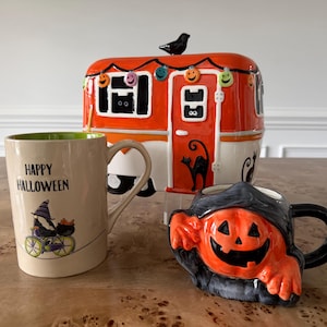 May include: A ceramic Halloween-themed camper cookie jar with a black bird on top, a beige mug with a witch on a bicycle and the text Happy Halloween, and a sculpted mug shaped like a grinning orange pumpkin with a black hood and handle.