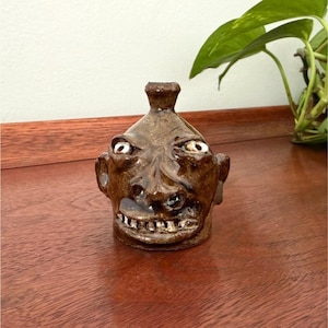 May include: A brown glazed ceramic face jug featuring a sculpted human-like head with prominent ears, a wide nose, and a mouth showing individual teeth. The top of the head has a small, narrow spout opening, and the surface has a glossy finish.