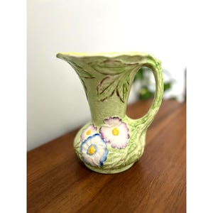 May include: A light green ceramic pitcher with a handle and a flared top. The pitcher is decorated with embossed leaves and three flowers with blue, white, and purple petals. The interior is yellow.