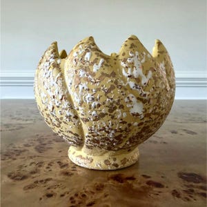 May include: A yellow ceramic vase with a unique, petal-like design. The vase is speckled with white and gold accents, and sits on a small, rounded base. The vase has a textured surface and a wide opening.