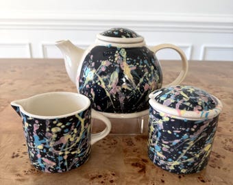 May include: A ceramic tea set consisting of a teapot, a creamer, and a lidded sugar bowl. Each piece features a glossy black base with a vibrant splatter pattern in shades of yellow, pink, and light blue. The teapot has a cream-colored spout and handle.