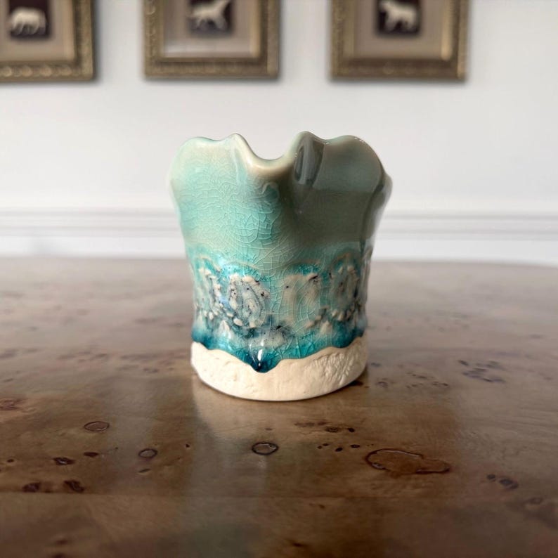 May include: A small, handcrafted ceramic vase with a unique, textured design. The vase features a turquoise and teal glaze with a white base. The rim has a wavy shape. The vase is displayed on a wooden surface.