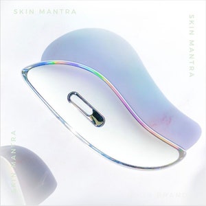 May include: A white and silver beauty tool with a curved design and a metallic accent. The tool has a light purple top and the text "SKIN MANTRA" is visible in the image.