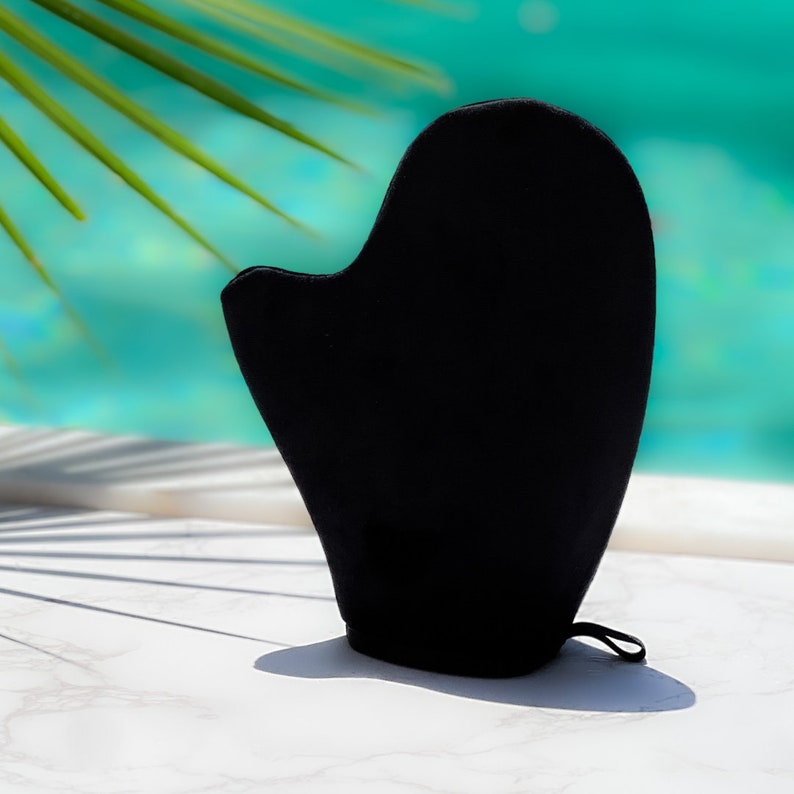 Pode incluir: Uma luva preta para autobronzeamento, em forma de m&atilde;o, com uma argola para pendurar. A luva &eacute; feita de um material macio e aveludado. O fundo apresenta uma vista desfocada de uma piscina e folhas de palmeira.