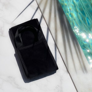 Pode incluir: Uma bolsa de veludo preta com uma al&ccedil;a, repousando sobre uma superf&iacute;cie de m&aacute;rmore. A bolsa &eacute; retangular com uma pequena al&ccedil;a na parte superior. O fundo inclui uma piscina e sombras.