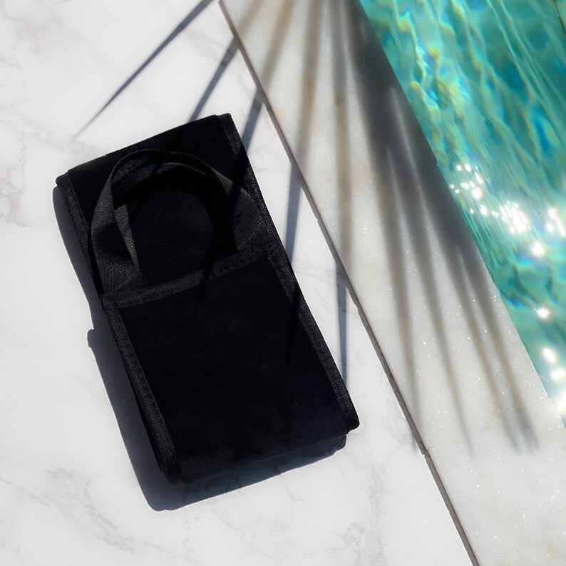 Pode incluir: Uma bolsa de veludo preta com uma al&ccedil;a, repousando sobre uma superf&iacute;cie de m&aacute;rmore. A bolsa &eacute; retangular com uma pequena al&ccedil;a na parte superior. O fundo inclui uma piscina e sombras.