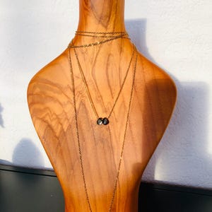 May include: Three gold-coloured necklaces with dark grey gemstone pendants are displayed on a wooden bust. The bust is light brown and has a smooth finish. The necklaces are draped around the bust, showcasing the different lengths and styles.
