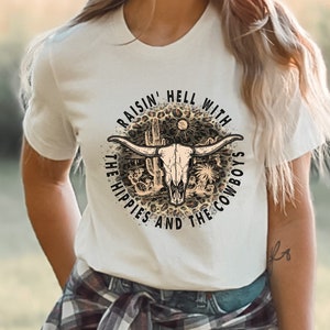 May include: White t-shirt with a graphic print of a skull with horns and the text "Raisin' Hell With The Hippies And The Cowboys".