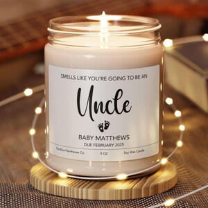 May include: A lit soy wax candle in a clear glass jar, with a white label that reads "Smells like you're going to be an Uncle." The label also includes the text "Baby Matthews Due February 2025." The candle sits on a wooden coaster.