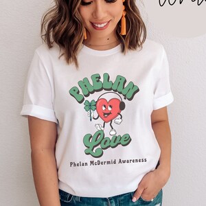 Può includere: Maglietta bianca con una grafica verde e rossa di un cuore con cuffie e un trifoglio a quattro foglie. Il testo "Phelan Love" è scritto in verde sotto la grafica. Il testo "Phelan McDermid Awareness" è scritto in nero sotto la grafica.