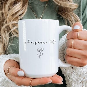Peut inclure: Mug en céramique blanche avec l'inscription "chapter 40" dans une police manuscrite, accompagnée d'une petite illustration florale. La tasse a une grande anse et est tenue par une personne aux cheveux blonds et portant un pull blanc.