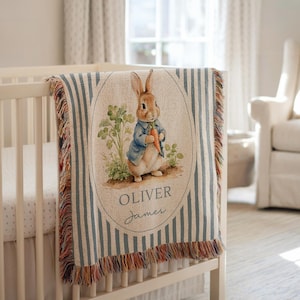 May include: A woven throw blanket featuring a blue and white striped pattern with a central oval illustration of a rabbit wearing a blue jacket and holding a carrot. The text "OLIVER" and "James" is printed below the rabbit illustration.