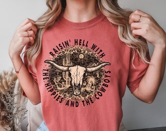 Könnte beinhalten: Ein rotes T-Shirt mit einem Grafikdruck eines Kuhschädels mit Hörnern, Kakteen und dem Text "Raisin' Hell With The Hippies And The Cowboys".