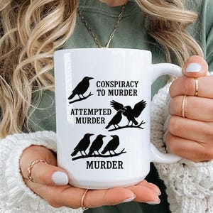 Puede incluir: Taza de cerámica blanca con ilustraciones de cuervos negros y las frases "Conspiracy to Murder", "Attempted Murder" y "Murder". La taza tiene un asa grande.