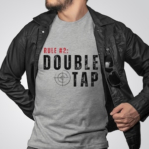 May include: A gray t-shirt with the text "RULE #2: DOUBLE TAP" in black and red. A target symbol is centered below the text.