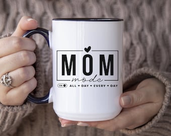 May include: White ceramic coffee mug with a black rim and handle. The mug has a black graphic that says "MOM mode" with a heart above the word "MOM" and the text "ON ALL DAY EVERY DAY" below.