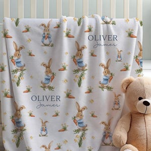 May include: A white baby blanket with a repeating pattern of watercolor bunnies in blue jackets holding carrots, with the name "OLIVER James" printed on it. A beige teddy bear sits next to the blanket in a white crib.