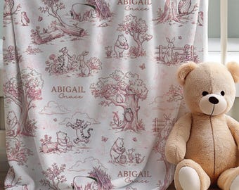 May include: A white baby blanket with a pink toile pattern featuring Winnie the Pooh characters, trees, and the name "Abigail Grace." A beige teddy bear sits next to the blanket in a white crib.