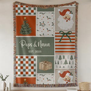 Può includere: Una coperta a tema natalizio con un design patchwork. La coperta presenta quadrati con un albero di Natale, Babbo Natale, agrifoglio e una scatola regalo. Il testo "Pops & Nana EST. 2025" è visualizzato su uno sfondo verde. La coperta ha un bordo sfrangiato.