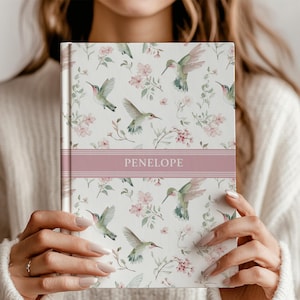 Pode incluir: Um diário personalizado com um padrão floral e beija-flores. A capa apresenta um fundo branco com flores rosa e beija-flores verdes. Uma faixa rosa no centro exibe o nome "Penelope" em texto branco. O diário é segurado por uma pessoa.