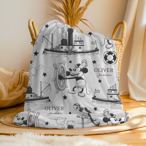 May include: A white fleece blanket featuring a black and white nautical pattern with illustrations of steamboats, life preservers, stars, and Mickey Mouse at a ship's wheel. The text "OLIVER James" is printed in a script and serif font combination.