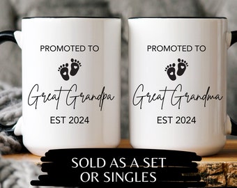 May include: Two white ceramic mugs with black rims and handles. The mugs have black text that reads "Promoted to Great Grandpa EST 2024" and "Promoted to Great Grandma EST 2024". Each mug features a black footprint with a heart in the center. The mugs are displayed on a wooden surface with a black banner that reads "Sold as a set or singles."