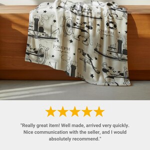 May include: A cream-coloured fleece blanket featuring a repeating black and white pattern of a cartoon mouse steering a steamboat, along with small stars and the printed text "JOSEPH Cooper". The design includes illustrations of a paddle steamer boat.