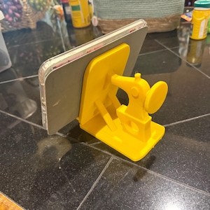 May include: A bright yellow 3D-printed plastic stand shaped like a vintage sewing machine, designed to hold a smartphone or tablet. The device is positioned upright against the flat back support of the stand, which features a base and a decorative arm.
