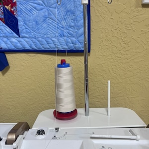 May include: A white sewing machine thread spool is on a white thread stand with a chrome arm. The thread stand is attached to a white sewing machine.