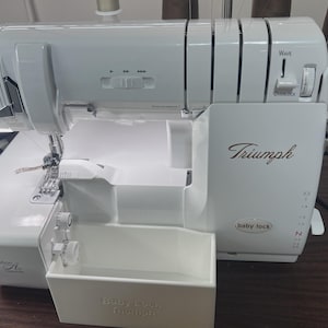 May include: A white Baby Lock Triumph serger sewing machine. The machine has a built-in waste bin and features the Triumph logo in a gold script. The machine has several dials and buttons for adjusting settings. The machine is on a wooden surface.