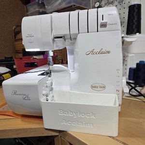 Puede incluir: Máquina de coser overlock Baby Lock Acclaim blanca. La máquina tiene el texto "Acclaim" y "Baby Lock" en la parte delantera. La máquina también tiene el texto "Revolution Air Threading" en el lateral.