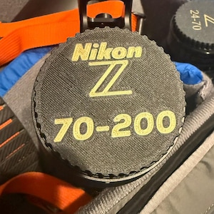 Puede incluir: Una tapa de lente de cámara negra con letras doradas que dicen "Nikon Z 70-200".