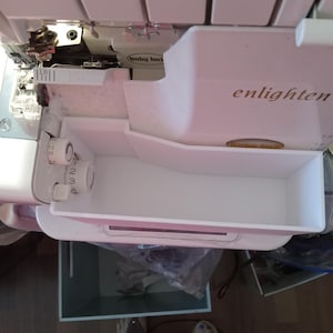 Könnte beinhalten: Weiße Nähmaschine mit einem weißen Plastik-Aufbewahrungskasten. Die Maschine hat ein Etikett mit der Aufschrift "Baby Lock" und "enlighten".