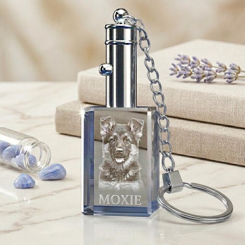 Puede incluir: Un llavero de cristal transparente con un retrato de perro pastor alem&aacute;n y el nombre "MOXIE" grabado. El llavero tiene una parte superior y una cadena de metal plateado, y un llavero. El fondo incluye una peque&ntilde;a botella de vidrio con piedras azules.