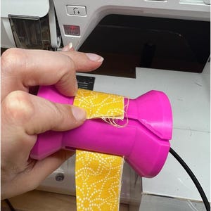 Puede incluir: Un enrollador de tela rosa con una tela amarilla con lunares blancos que se enrolla alrededor. El enrollador está siendo sostenido por una mano frente a una máquina de coser blanca.