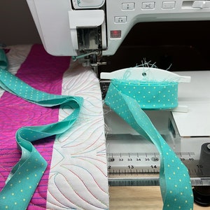 May include: A close-up of a sewing machine with a spool of turquoise fabric with white polka dots. The fabric is being fed into the machine and is being used to bind a quilt with pink and white fabric. The quilt has a white background with a pattern of curved lines in different colors.