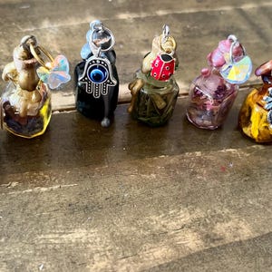 Può includere: Una collezione di dieci piccole bottiglie di vetro con vari ciondoli e decorazioni. Le bottiglie sono riempite con liquidi e cristalli di diversi colori. I ciondoli includono una farfalla, una mano di hamsa, una coccinella e un fiore.