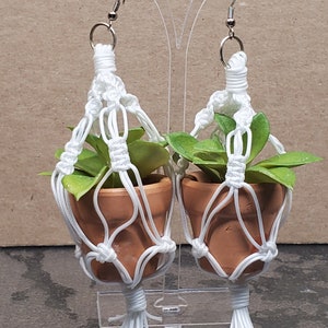 May include: A pair of white macrame plant hangers with small terracotta pots and green plants. The hangers are attached to silver earring hooks.