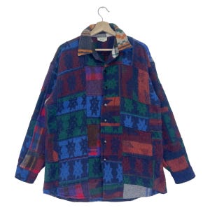 Op de afbeelding: Een lang mouw overhemd met knopen en een patchwork ontwerp. Het overhemd is gemaakt van een zachte, pluche stof en heeft verschillende kleuren, waaronder blauw, rood, groen en bruin. Het overhemd heeft een kraag en een knoopsluiting aan de voorkant.